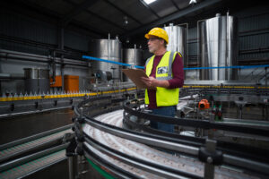 Arbeiter mit gelbem Schutzhelm und Warnweste kontrolliert Produktionsanlage in einer Fabrikhalle.