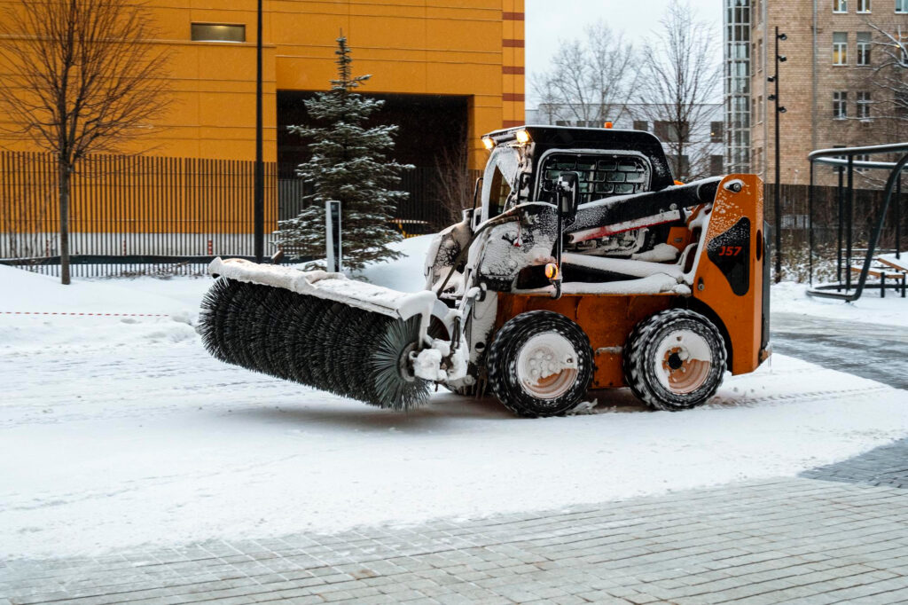 Kompaktlader mit rotierender Schneebürste räumt Schnee auf einer Straße vor einem Gebäude.