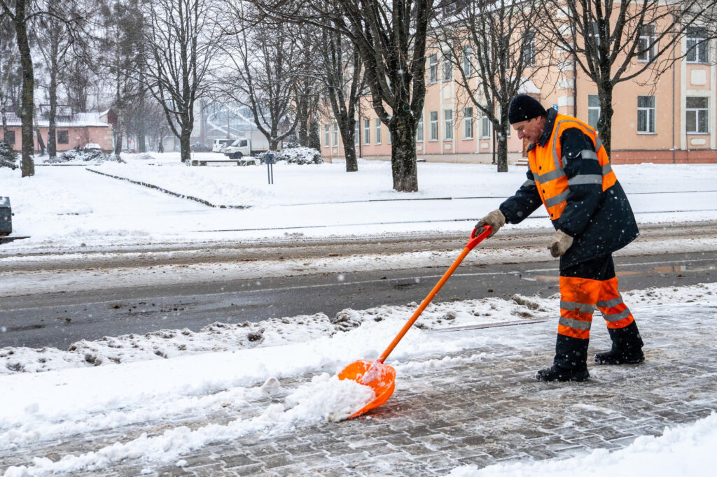 Mitarbeiter in orangefarbener Warnkleidung räumt Schnee von einem Gehweg mit einer Schneeschaufel.